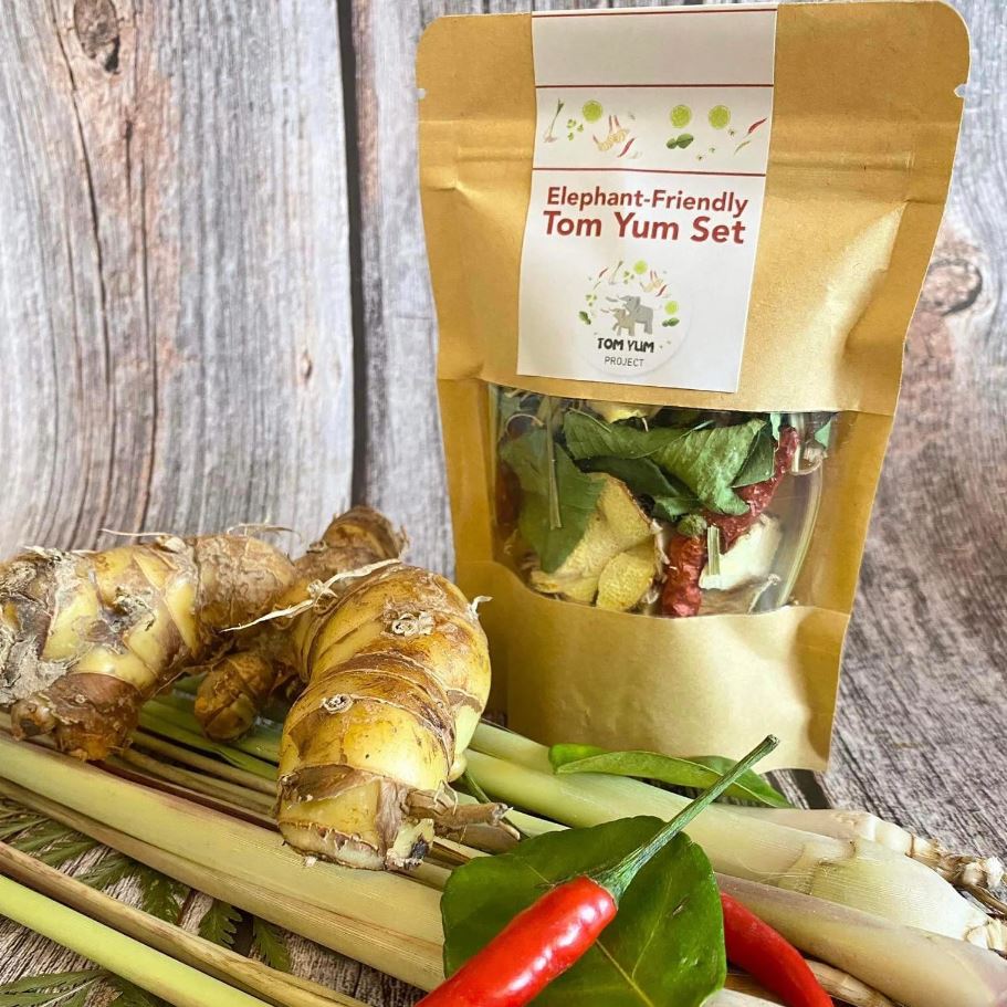A package of elephant-friendly Tom Yum soup ingredients surrounded by fresh ginger, lemongrass, and chili peppers.